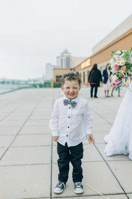 A little boy wearing a bow tie stands on a sidewalk, smiling and looking towards the camera.