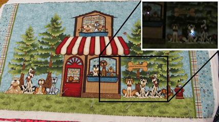 A quilt with a scene filled with dogs, a house, and trees. Some of the dogs’ collars are lit up with LEDs in the dark.