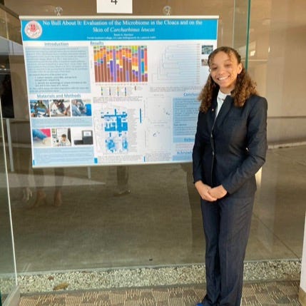 Raven stands beside a scientific research poster displayed on a glass wall. The poster includes charts, graphs, diagrams, and text sections describing a research project, with the presenter positioned to the right of the poster. Raven&rsquo;s long, curly, brown hair is pulled back into a half-up half-down style. She looks very professional and is wearing a black suit with a white dress shirt underneath.