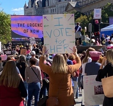 Sign reading “I vote for her” with arrows pointing to the crowd.