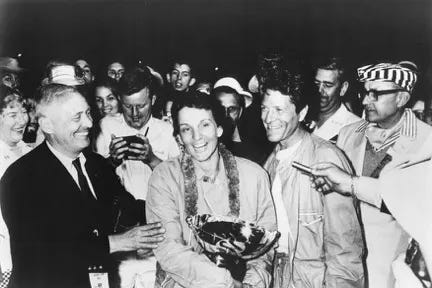In this black-and-white photo, a crowd of people smile and take photos as a smiling woman holds a trophy. One her right, a man in a suit touches her arm; he seemingly just presented the trophy. On her left, her co-driver smiles wryly.