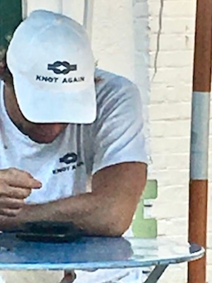 Color photo midshot image of a young tanned man seated at some outdoor table, with his arms bent staring down at what appears to be his phone, wearing a matching white ballcap hat and white short-sleeved polo-style shirt, both with the logo “KNOT AGAIN” printed in dark blue.