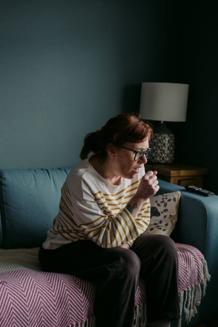 A woman sits by herself on her couch and coughs into her hand