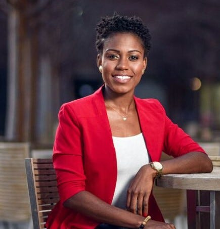 In this headshot, Dr. Keturah Adams sits at an outdoor table wearing a red blazer over a white shirt. Her hair is styled in a natural coiled short style. She is smiling at the camera.