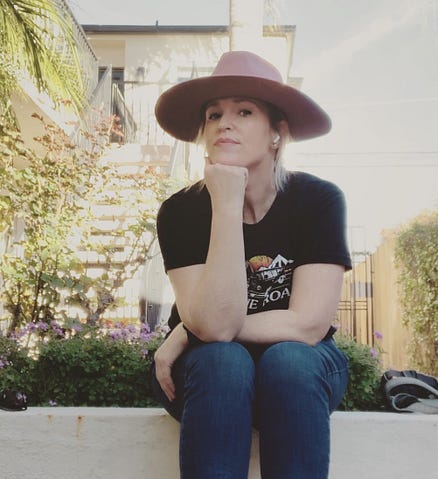 The author sits facing the camera. Her head resting on her hand. She wears a black t-shirt, jeans, and a large pink rancher hat.