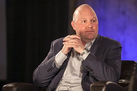 photograph of Marc Andreessen, sitting thoughfully with fingers interlaced, a bit of theatre scrim visible in the background, as if he is on a stage, perhaps participating in a panel discussion, his bald head domed and prominent, wearing a gray suit jacket with no tie, and a large, silver-colored wristwatch