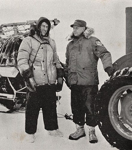 Black and white newspaper image of Hillary and Dufek standing behind a tractor at Scott Base. Both men are wearing thick jackets, mittens and have fur lined hoods.