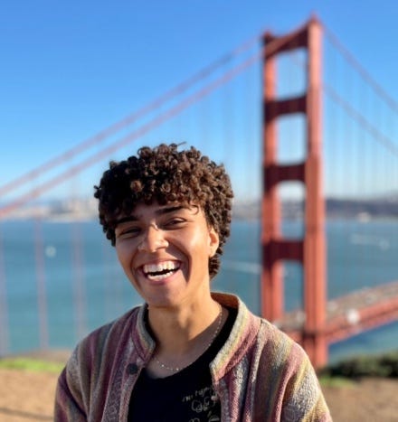 Morgan P. Vickers — a Black queer nonbinary person with brown skin and dark brown, short, curly hair — stands in the center of the frame, grinning cheesily. They’re wearing Black Uniqlo shirt with a gold Jean-Michel Basquiat design on the front, a gold chain, and a pink cotton jacket. They’re standing in front of the Golden Gate Bridge on a clear blue day.