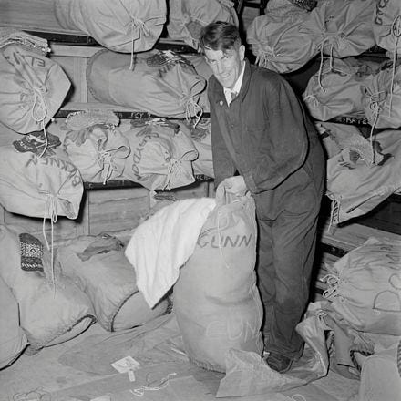 Black and white photograph of Sir Edmund Hillary in a warehouse with sacks stacked behind him. He is stuffing a white blanket into the sack and smiling at the photographer .