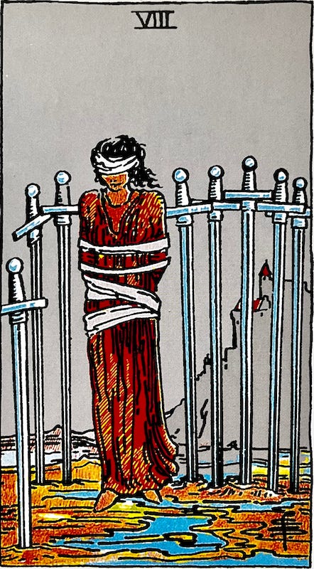 EIght of Swords — A woman stands trapped with eight swords around her. SHe is bound around her arms and body and in blindfolded.