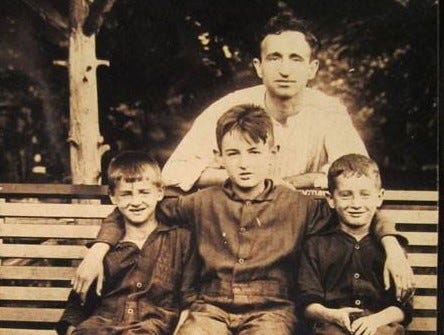 three young boys in front, twins to either side of the middle boy, who is perhaps two years older. A father stands in back. This is an old sepia tone photo.