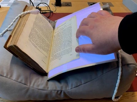 A reader consulting a printed book, which is supported open on a book cushion with a light sheet inserted underneath the right hand page.