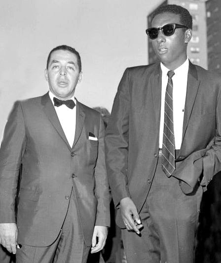 A picture of Rev. cleage and Stokely Carmichael with suits. On the left, Rev. Cleage wears a bow tie and on the right, Stokely Carmichael wears a tie. The Jefferies Housing Project is in the back.