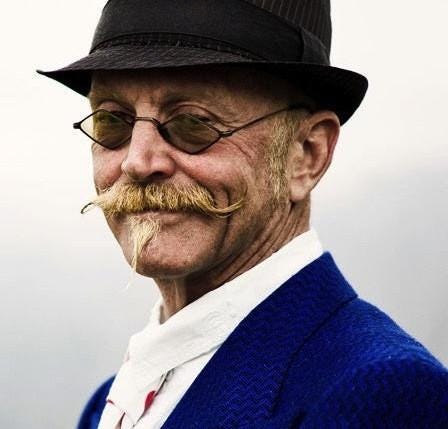 head and shoulder view of Gary in quirky hat, glasses, waxed handlebar moustache, and midnight blue suit.