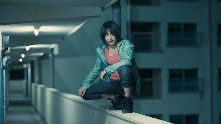 A woman wearing athletic clothing squats on top of a low wall in an apartment building. It’s nighttime.