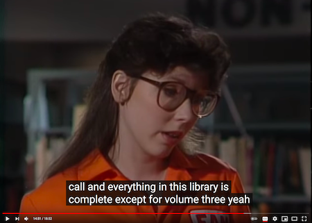 screenshot from video, woman in an orange jumpsuit and 80s style glasses “Everything in this library is complete…”