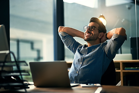 Man sitting at his desk, leaning back with his hands behind his head and a smile of confidence and no worries on his face.