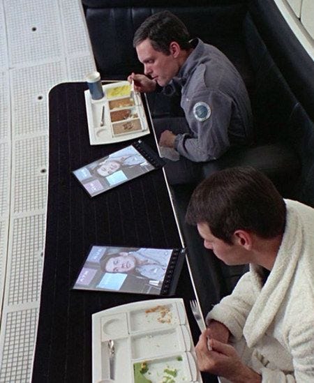 Cena do filme 2001 — Uma Odisseia no Espaço onde 2 homens almoçam enquanto olham duas telas que parecem tablets/ipads.