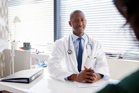 Doctor in a white lab coat and tie sitting at his desk smiling at his patient.