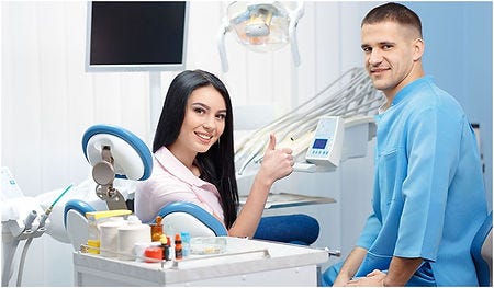 Picture of attractive female dental patient with long dark hair sitting in the dental chair giving a thumbs up to the camera and with a smil on her face. There is a smiling male dentist in blue scubs standing next to her also smiling at the camera.