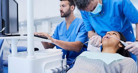 Picture of a dentist in blue scrubs doing a precedure on a patient with an assistant typing on a keyboard for a computer connected to stae of the art dental equipment.