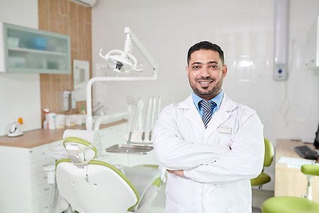 Picture of a Happy Confident Male Dentist or Orthodontist Standing in front of a dental chair and equipment with His Arms Folded and a smile on his face.