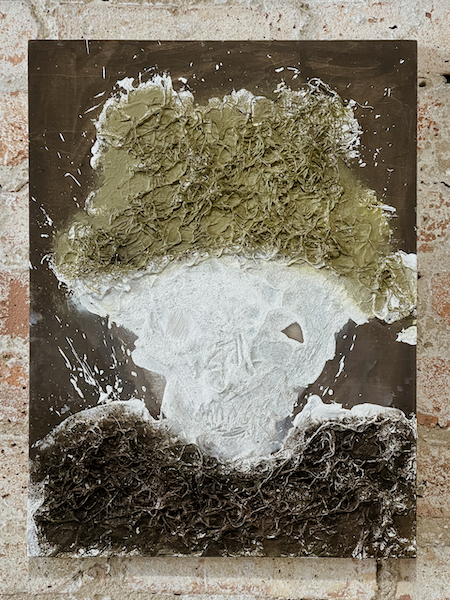 A mixed-media artwork by Alexis de Chaunac called “Fur Coat and Fur Hat” created in 2020. Inside the frame is a figurative body that looks like a ghostly figure wearing an olive-colored fur hat and a brown fur coat. The hat and coat are made using moss.