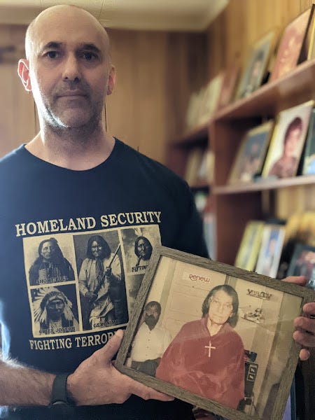 A Native American man visits his parents home and poses in front of a wall of family photos. He holds a framed portrait of his grandmother, who wears a cross necklace. His own shirt features five Indian chiefs and reads “Homeland Security, Fighting Terrorism Since 1492.”