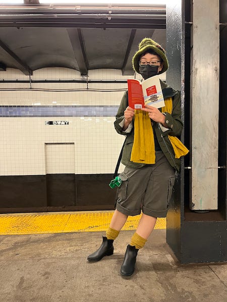 A photo of a person leaning against a pillar in the New York subway, dressed as Snufkin for Hallowe’en, wearing a face mask, reading The Summer Book.