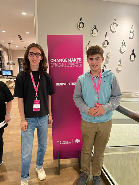Two young men standing in front of a vertical pink banner that says “Changemaker Challenge registration.” They are both wearing pink lanyard name tags and smiling.