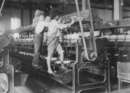 A black and white photo of barefoot child labourers working the yarn machines in the industrial revolution.