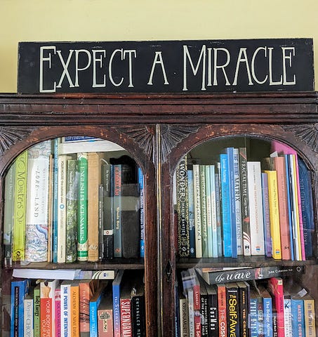 The author’s bookcase, with a sign over it reading: Expect a Miracle.