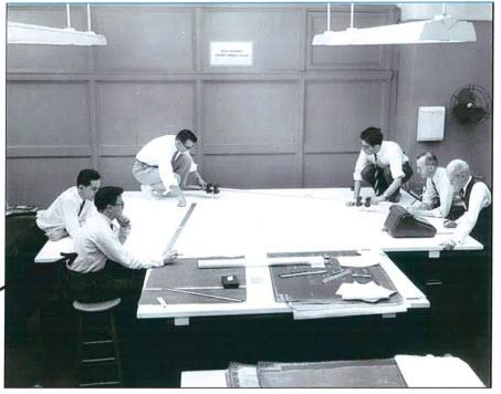 Black-and-white photo shows six men in office attire around a large table with blueprints laid out on it.