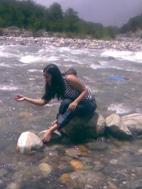 The author sits on a rock in a shallow stream as she lets go of some water she had scooped up in her right hand.