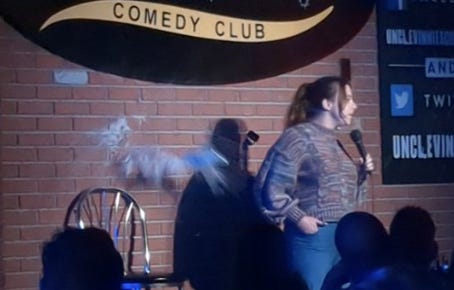Comedian Ariel Elias stands on stage at Uncle Vinnie’s Comedy Club in Point Pleasant Beach, New Jersey. The large comedy club logo hanging on the red brick wall behind her. A silver metal chair to her right, brown hair pulled back, and mic in hand. Just above the chair a full can of beer explodes against the brick wall.