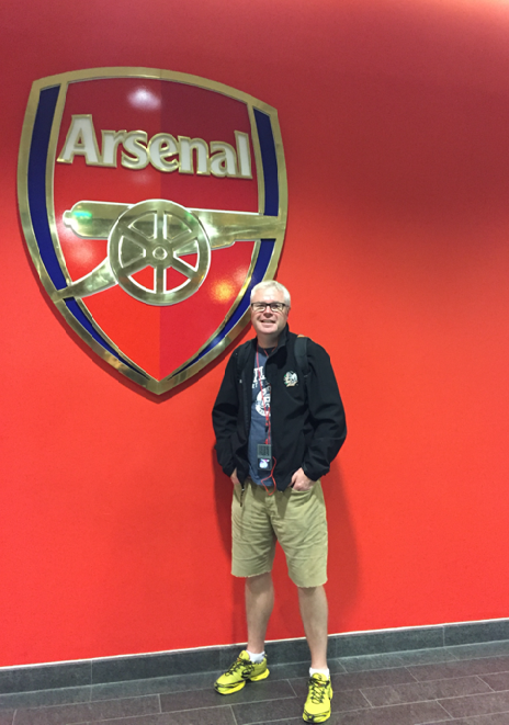 A man standing in front of a red wall with the Arsenal club logo on it.