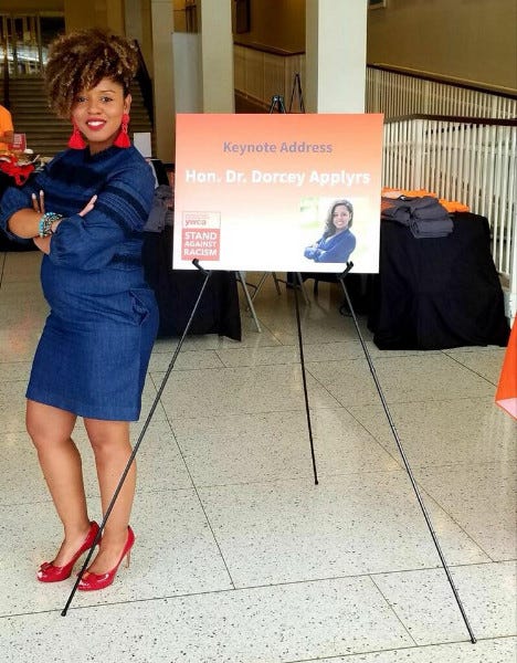 Dorcey Applyrs stands next to an easel displaying a poster for a speech she will give.