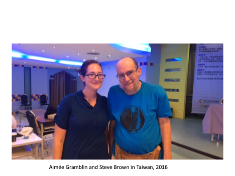 Standing at a conference in Taipei, Taiwan are Aimée Gramblin and Steve Brown. Aimée, Steve’s daughter and attendant, is wearing a dark blue shirt and has brown hair pulled back from her face and glasses. She is standing next to, and touching, Steve, a balding man with brown hair and beard and wearing glasses and a blue shirt with an unreadable picture on it.