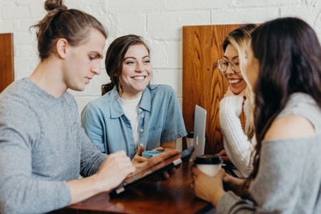 A group of four young adults are sitting close together at a coffee shop talking, smiling, and laughing