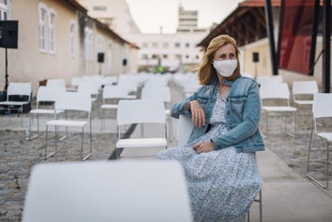 A woman wearing a cloth facemask sits alone outside. She is sitting in one of many chairs that have been arranged to accomodate social distancing.