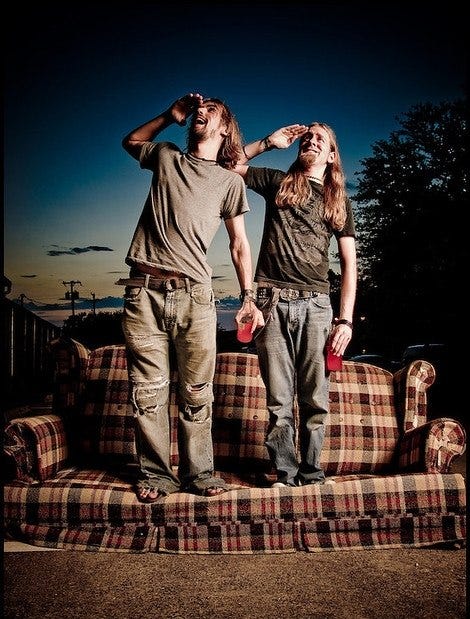 Two hippie looking men on a couch, saluting in a funny way