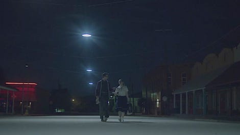 A man and a woman in 1950s attire walking along a street at night.