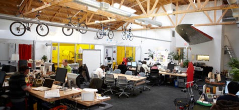 A picture of IDEO’s Palo Alto office around 2008. It is a colorful open-plan office with exposed beams and bicycles stored by hanging them from the roof, and and airplane wing jutting from one wall.