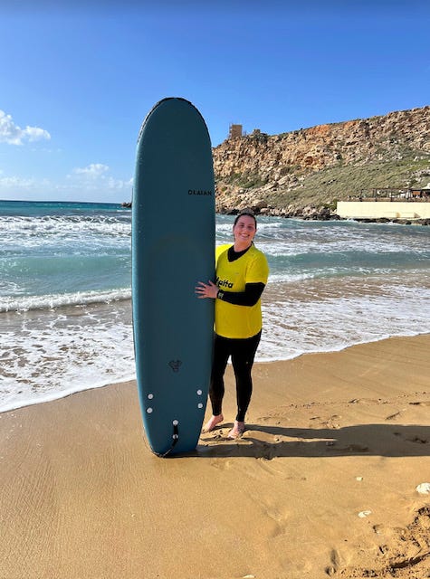 A girl in a yellow shirt and black wetsuit stands on a beach with a blue surfboard.