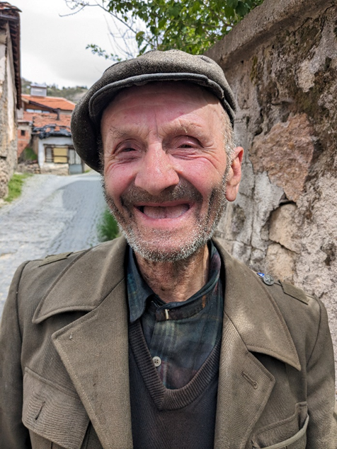 A smiling old man with a scruffy beard, no teeth, and tattered clothing
