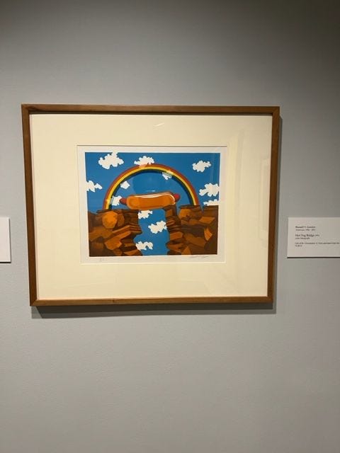 A painting on a gallery wall. The painting features a hotdog acting as a bridge between two cliffs. There is a rainbow above the hotdog bridge and white clouds in a blue sky. behind the rainbow.