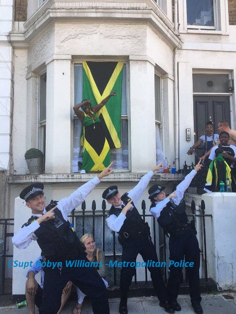 3 white police officers srand in front of a house adorned with the Jamaican flag. They strike Usain Bolt's lightening pose as they smile into the camera.