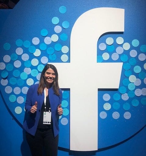 Laurie Keith stands in a blue jacket and black jeans in front of a giant Facebook logo with various blue dots.