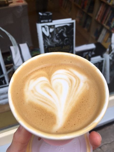 A flat white in a pink paper cup, with a bookshop window behind.