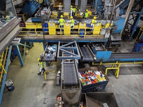 Workers at a recycling facility sort and separate recycled plastics into individually labeled yellow bins.